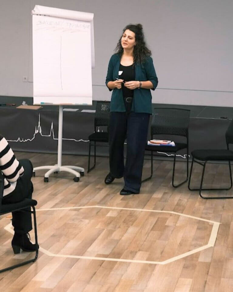 Woman presenter in a teal cardigan stands beside a flip chart, speaking to an audience in a training room.